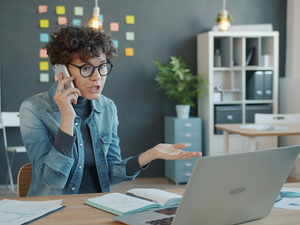 Angry young woman is speaking on mobile phone in office discussing problems at work gesturing expressing negative emotions. Youth and communication concept.