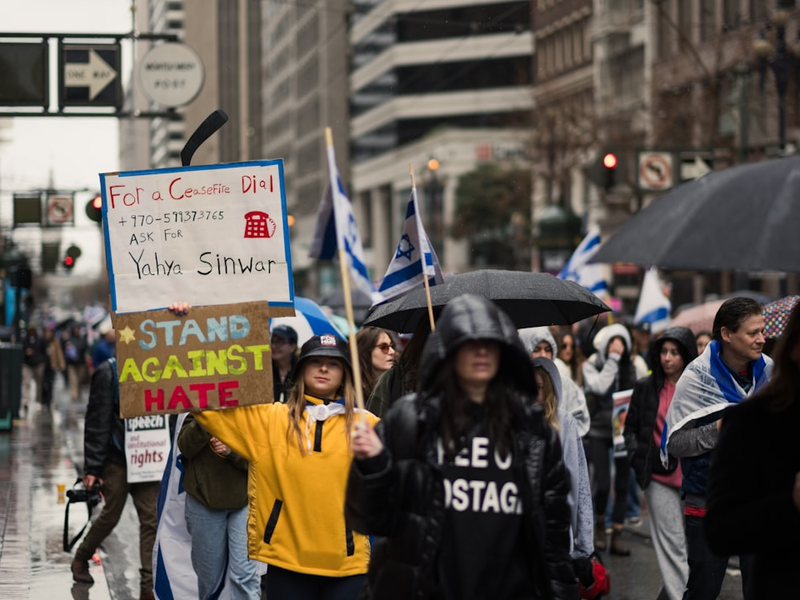 At the Unity Rally, a march against antisemitism held in San Francisco, an activist holds up a sign stating "STAND AGAINST HATE", while another holds up a sign state stating "FOR A CEASEFIRE ASK FOR YAHYA SINWAR" as a statement against Hamas efforts to block a durable, honest ceasefire. Other demonstrators hold up Israeli flags.