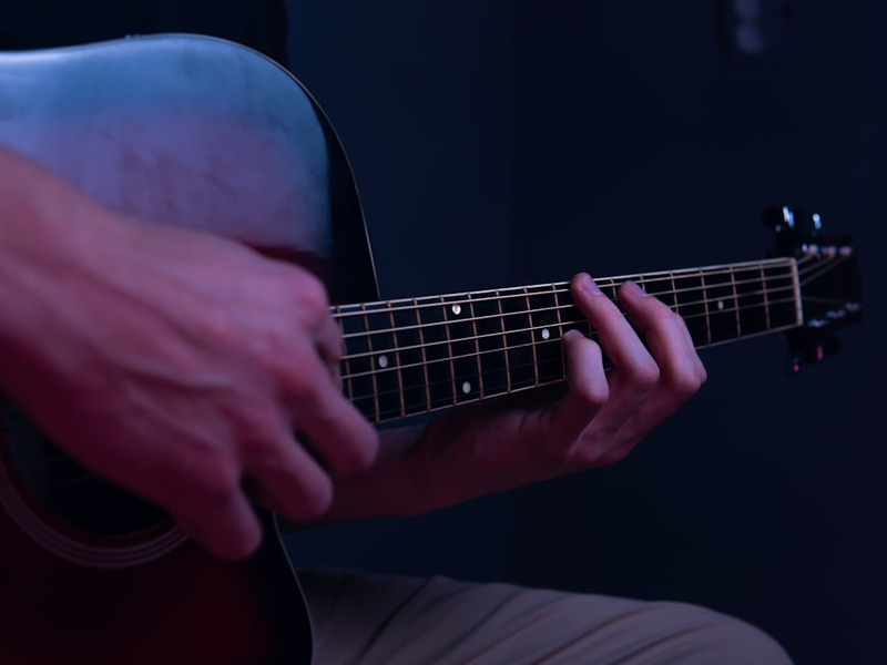 An adult man plays a red acoustic guitar on a dark background. Strong blond guy. Guitar lessons. Performance of the guitarist on the blue-red stage. Portrait of beautiful boy with a musical instrument