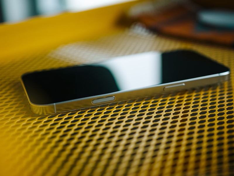 A close-up of an iPhone 16 Pro Max resting on a textured yellow surface. The reflection of light on the screen adds a sleek, modern feel to the image, while the geometric pattern of the background creates a striking contrast with the phone’s smooth edges. The composition emphasizes the minimal design and premium materials of the device.