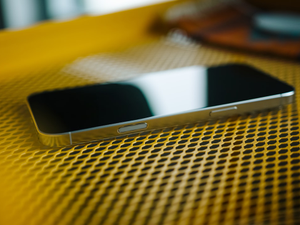 A close-up of an iPhone 16 Pro Max resting on a textured yellow surface. The reflection of light on the screen adds a sleek, modern feel to the image, while the geometric pattern of the background creates a striking contrast with the phone’s smooth edges. The composition emphasizes the minimal design and premium materials of the device.