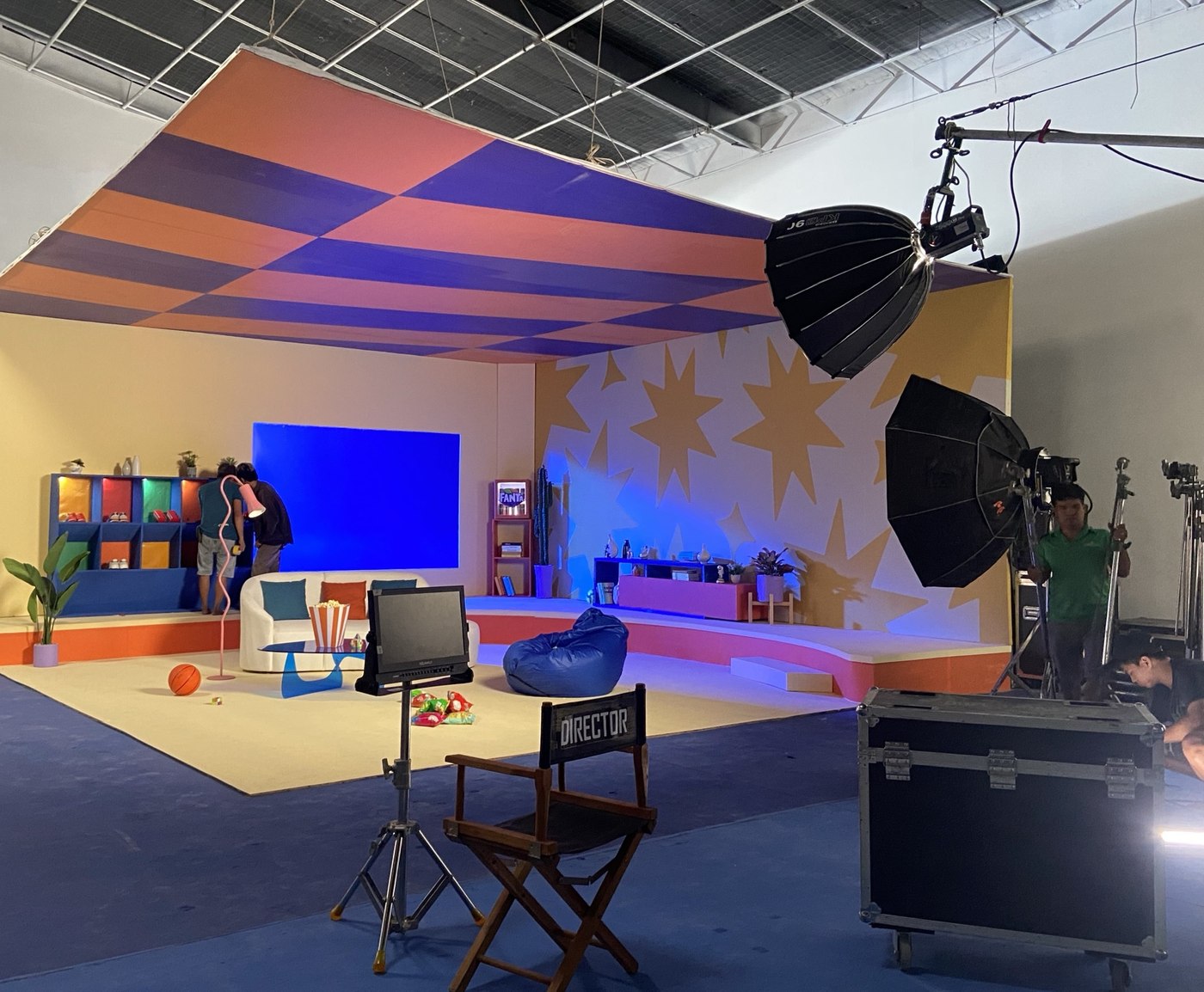 A colourful set built in a studio for a Fanta film, with a checked orange and purple ceiling, a star-patterned wall, a blue panel and soft lights on stands, seen from beside the director’s chair