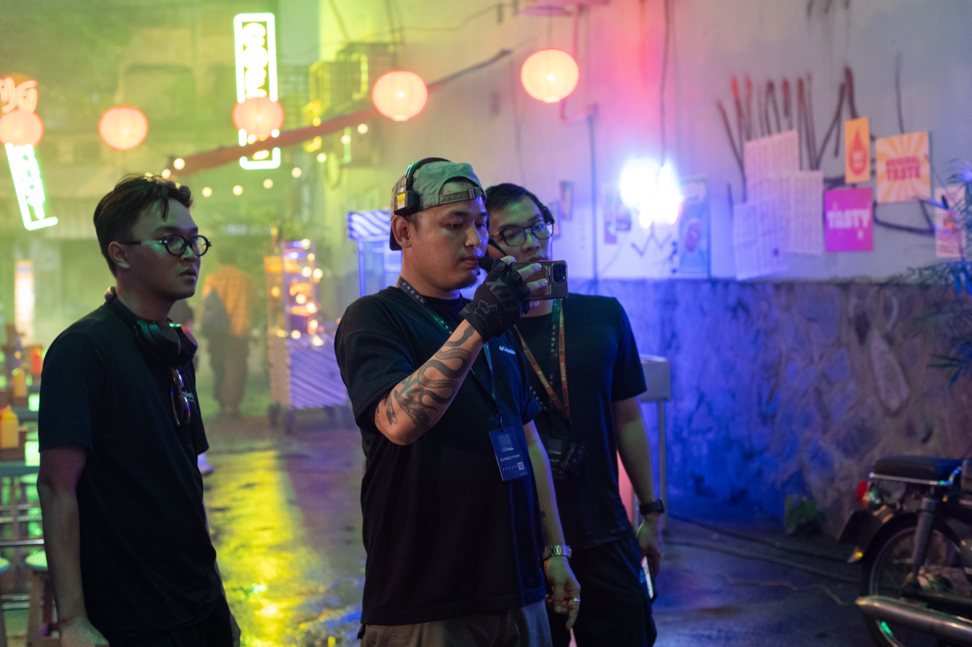 Three crew members looking at a frame on a phone in a wet, neon-lit alley with lanterns and haze during a Samsung Galaxy S24 night shoot