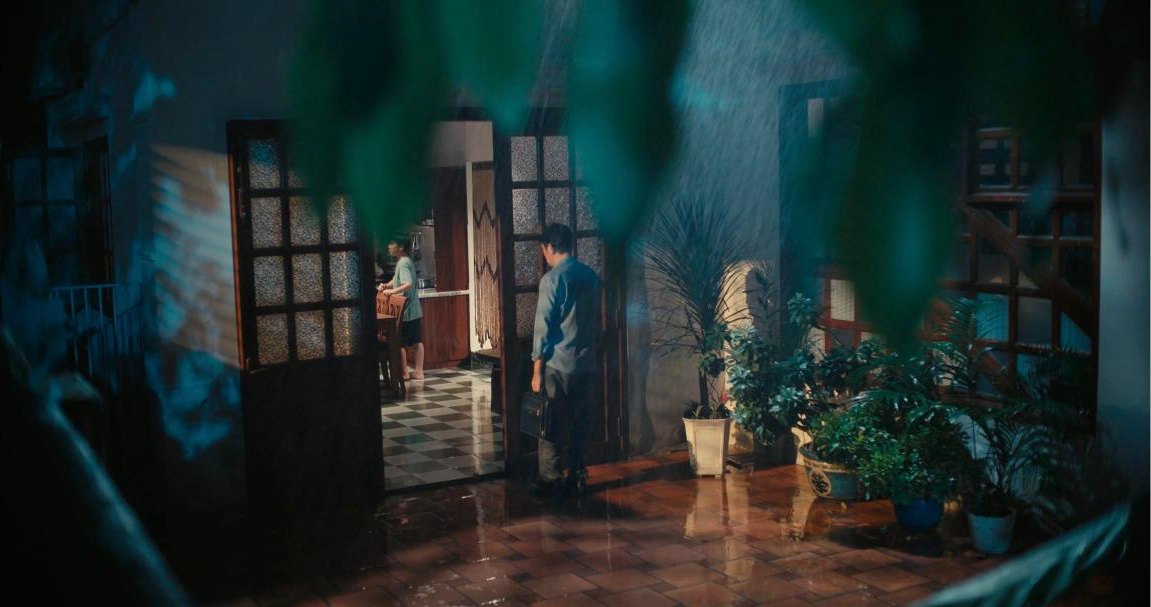 Night courtyard in heavy rain, a man with a briefcase at an open door and the wet tiled floor reflecting the light, from the short film Thương Phổi