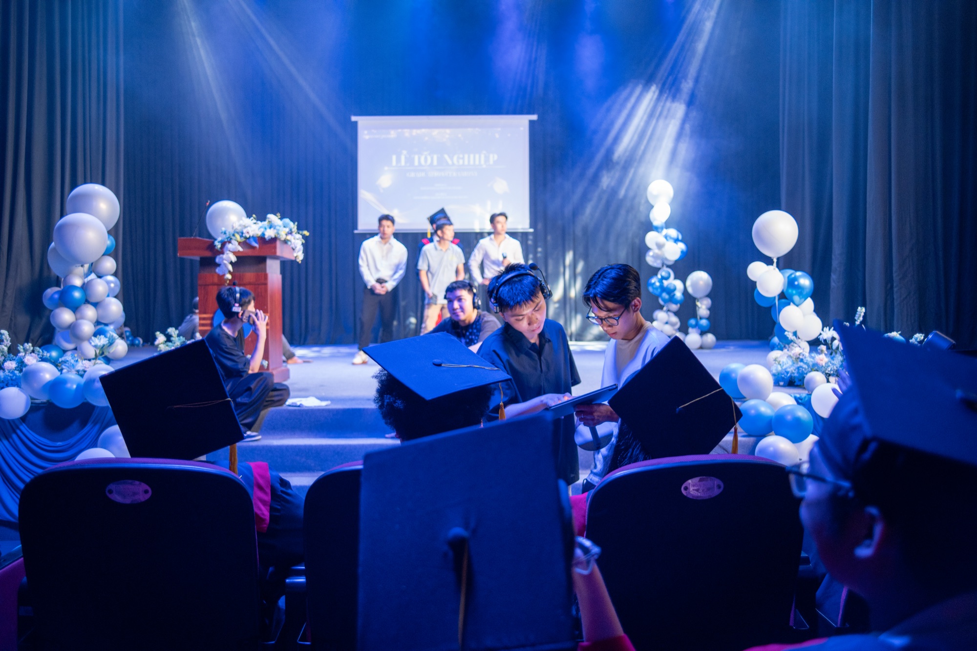Extras in graduation caps seated in an auditorium facing a stage dressed for a graduation, with crew on the stage, during a Samsung Galaxy A56 shoot