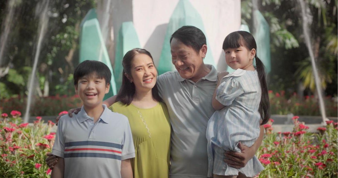 A family of four smiling in a park, the father holding his young daughter, from the short film Thương Phổi