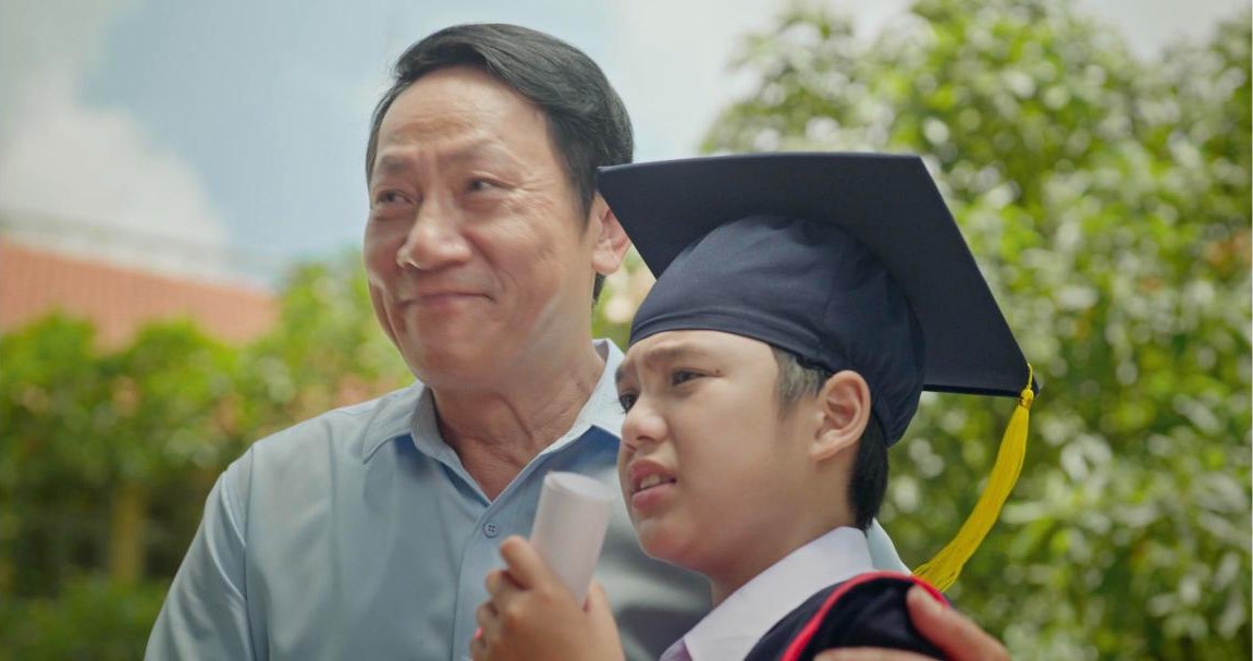 A father with his arm around his son in graduation cap and gown, both moved, from the short film Thương Phổi