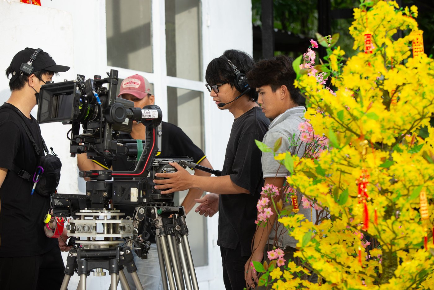 Camera crew in headsets setting up a cinema camera on a tripod beside a flowering apricot tree hung with red Tết decorations, on a Fanta shoot