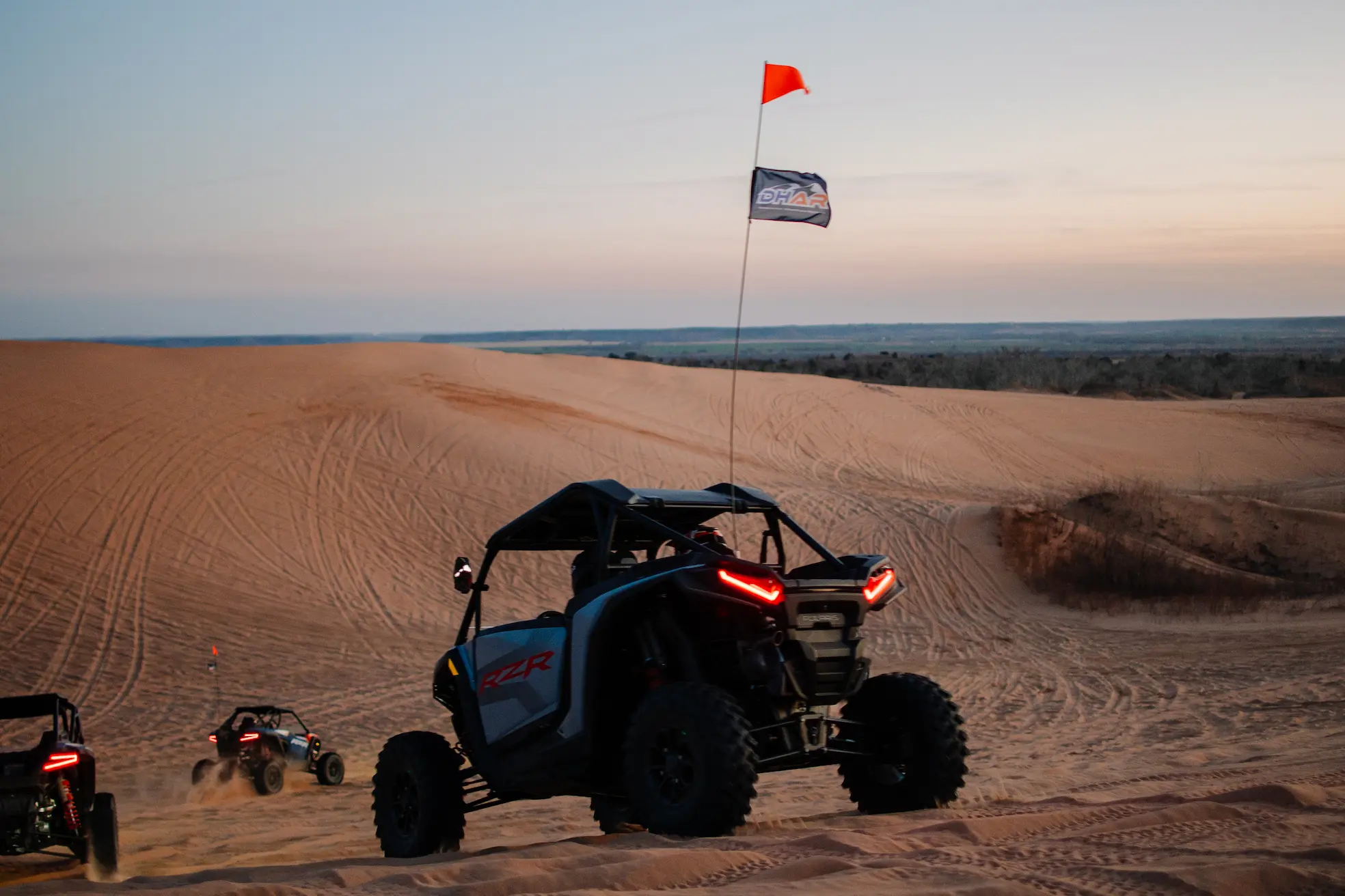 A dark gray Polaris RZR side-by-side cresting a sand dune at dusk, flying a Duner's Hut Adventure Rentals (DHAR) flag, with two other UTVs trailing below at Little Sahara State Park in Waynoka, Oklahoma.