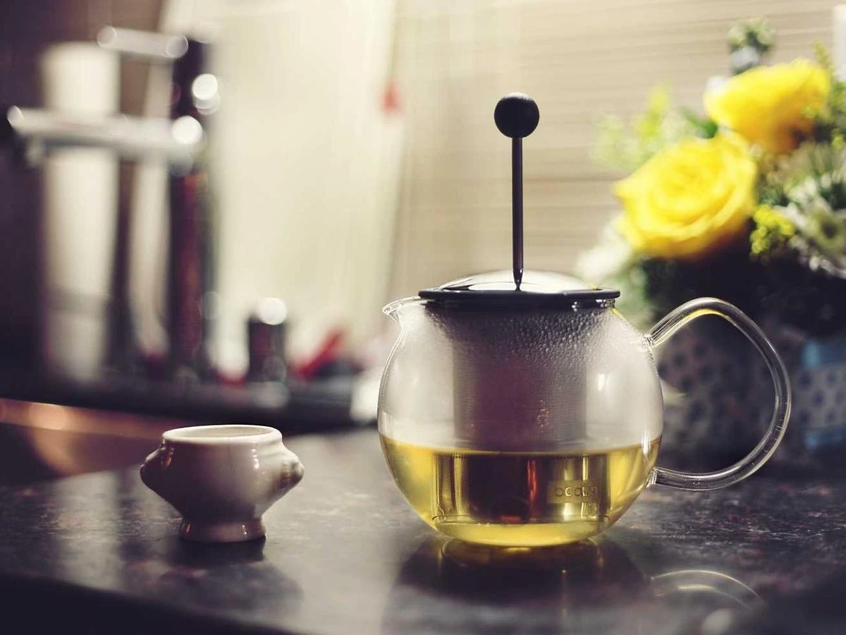 A glass tea press of golden tea beside yellow flowers