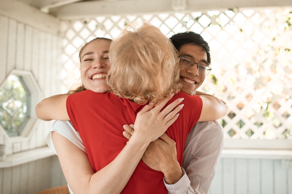 Three people smiling in a group hug in a bright sunlit room