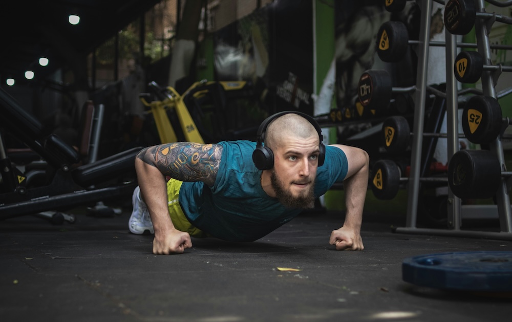 A man wearing headphones doing a push-up on a gym floor, surrounded by weight racks