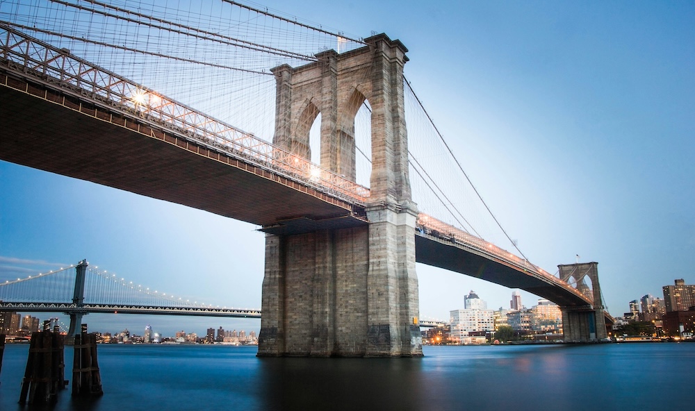 The Brooklyn Bridge at dusk seen from the water, with the Manhattan Bridge and the Brooklyn waterfront behind it