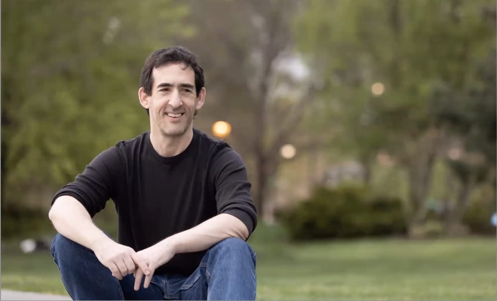 Mark, a male cuddler in NYC, sitting outdoors in a park and smiling at the camera