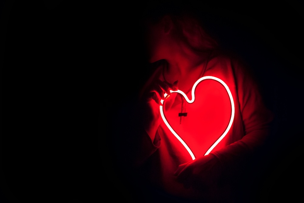 A person holding a glowing red neon heart against their chest in a dark room