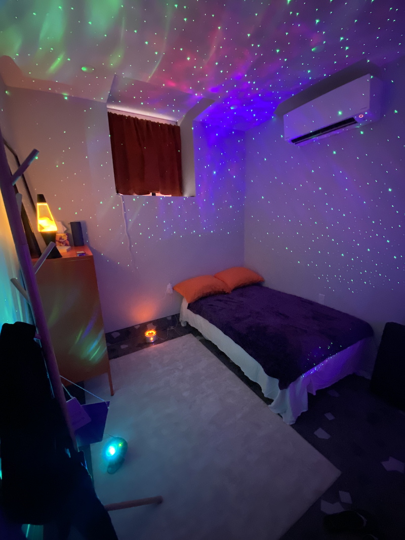 Inside the Embrace Club cuddle studio: a low bed with orange pillows and a deep purple blanket, a lava lamp glowing on a dresser, and a star projector washing the walls and ceiling in green and violet light