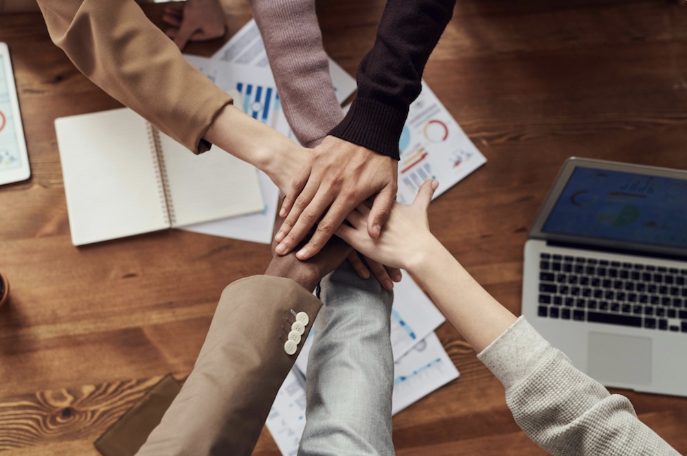 Several people stacking their hands together over a desk covered with notes and charts