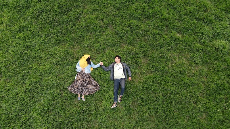 Overhead view of two people lying on their backs in tall green grass, holding hands and smiling at each other