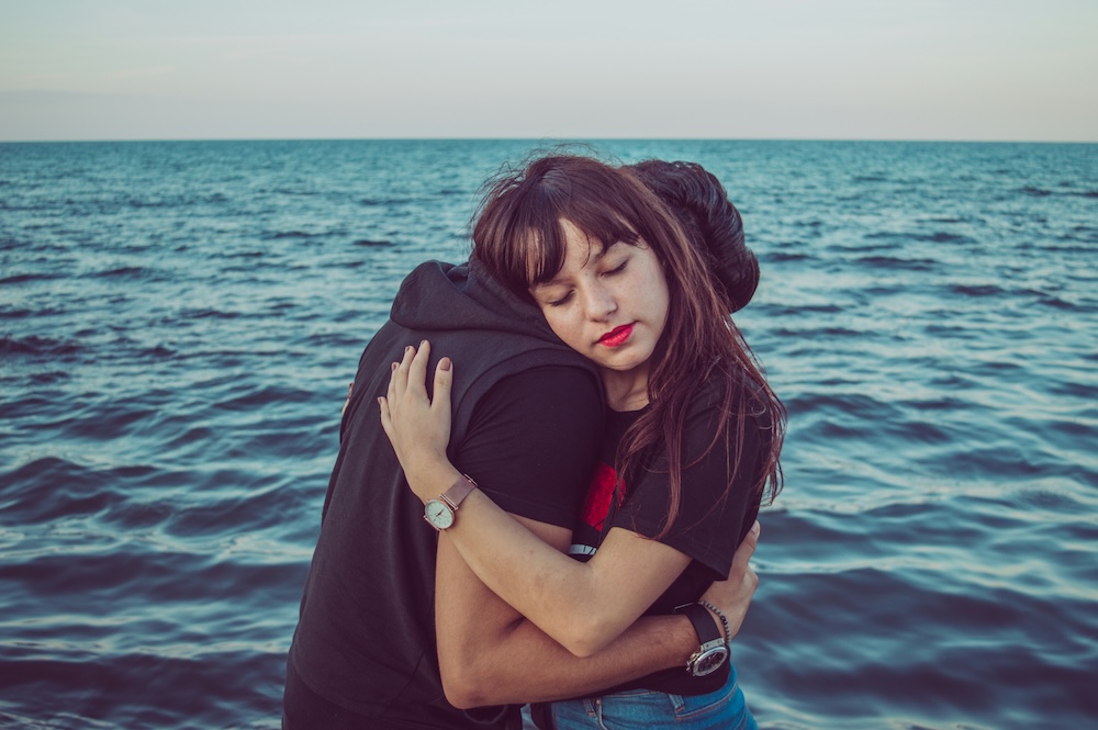 Two people holding each other in a close embrace at the water's edge, with the sea behind them