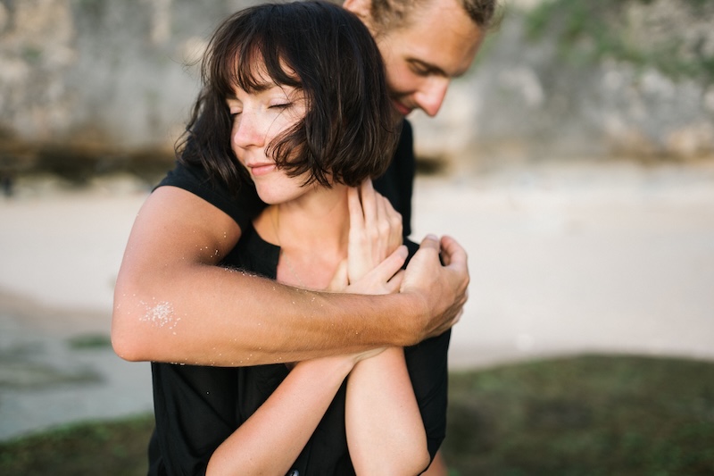 A man hugging a woman from behind on a sandy beach, her eyes closed and smiling as she holds his forearm