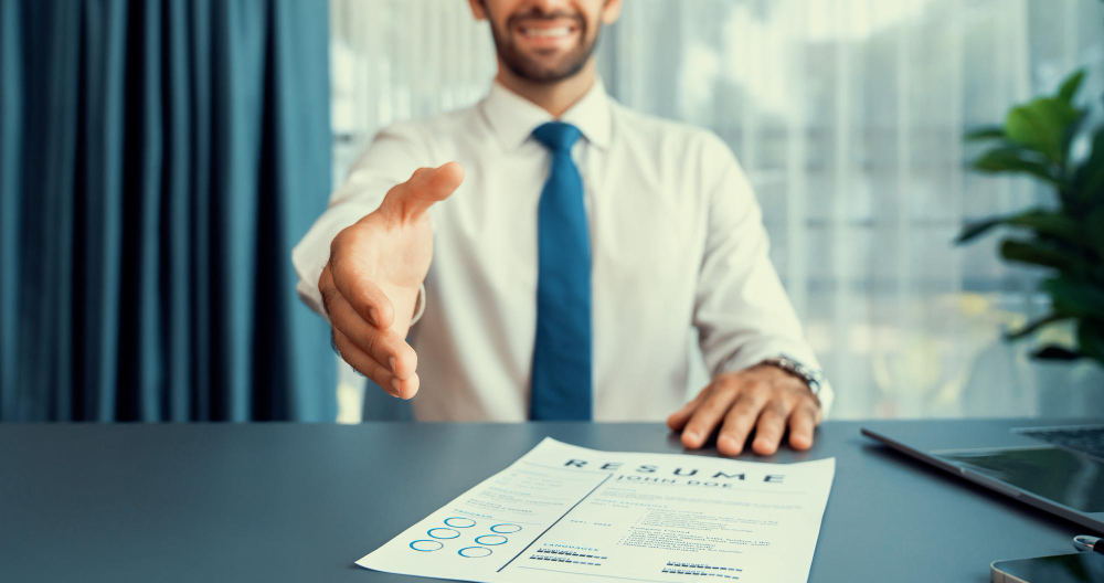 Angajator care întinde mâna unui candidat la interviu, cu un CV pe birou