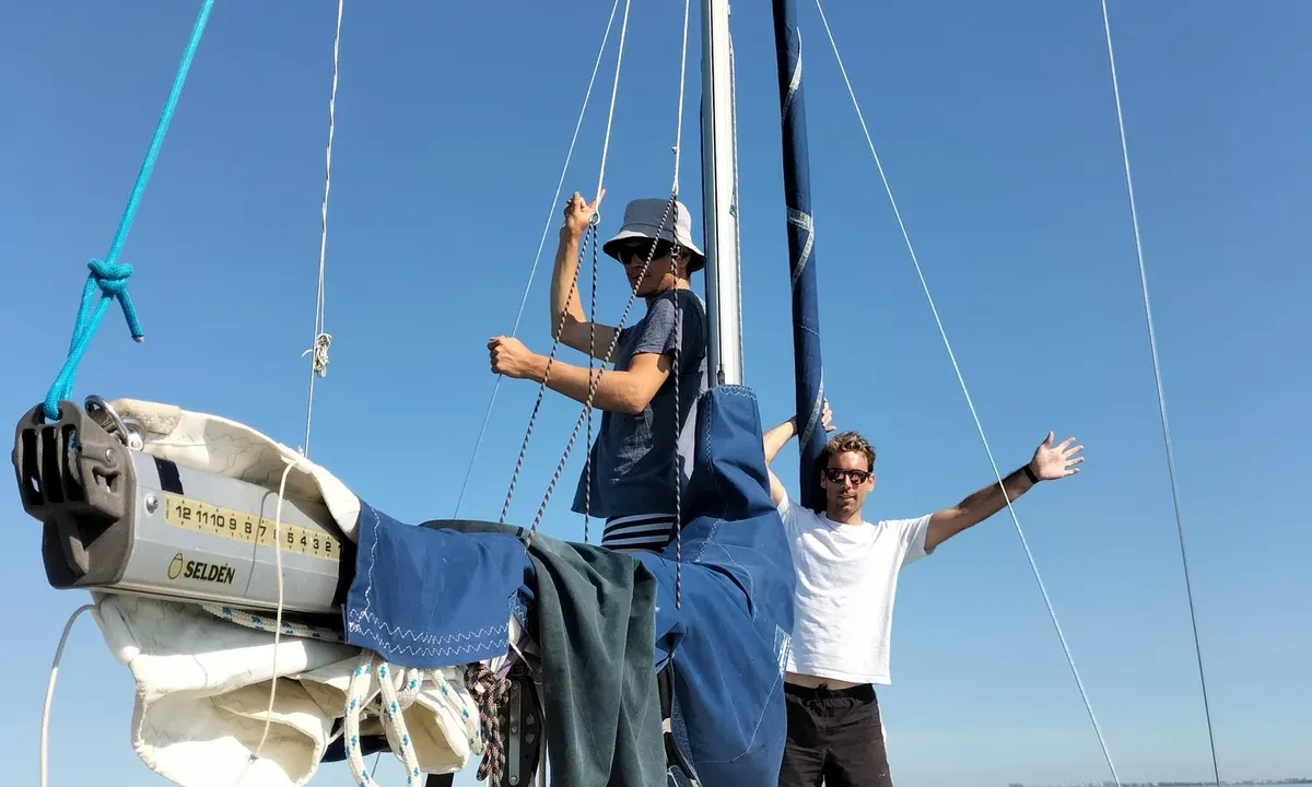 Two crew members on the deck of a sailboat, arms raised in front of the mast