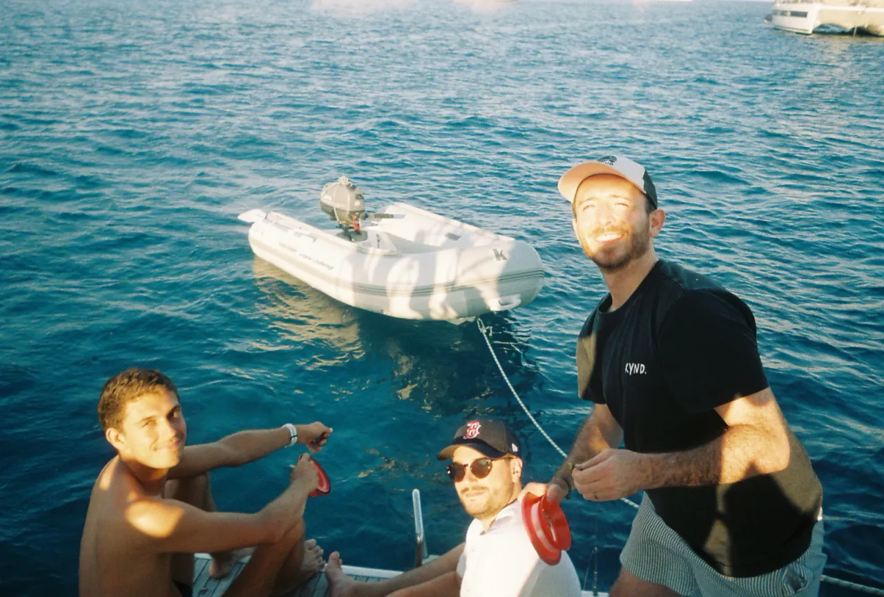 Three smiling crew members on the deck of a sailboat, with a tender behind them