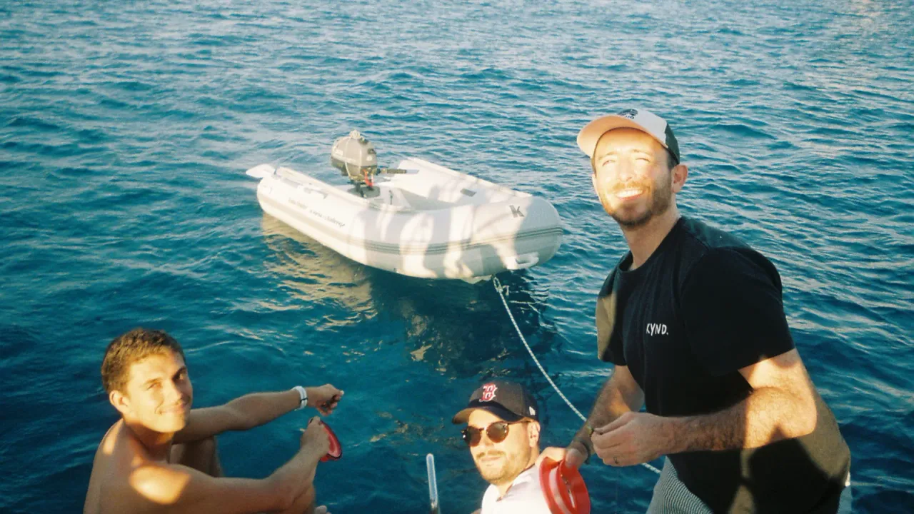 Three smiling crew members on the deck of a sailboat, with a dinghy behind them