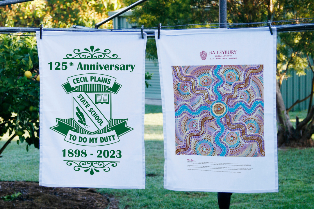 Two custom tea towels hanging outdoors – one celebrating a 125th anniversary with a school crest, the other showcasing colourful Aboriginal-inspired artwork.