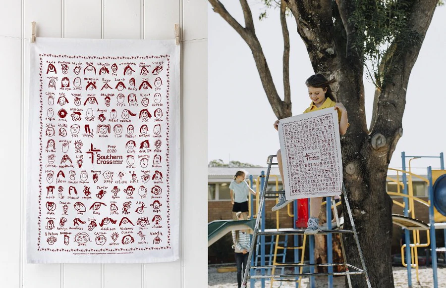 Custom printed school tea towel featuring children’s hand-drawn self-portraits from Southern Cross Catholic College, held by a student outdoors on school playground equipment during a tea towel fundraising event.