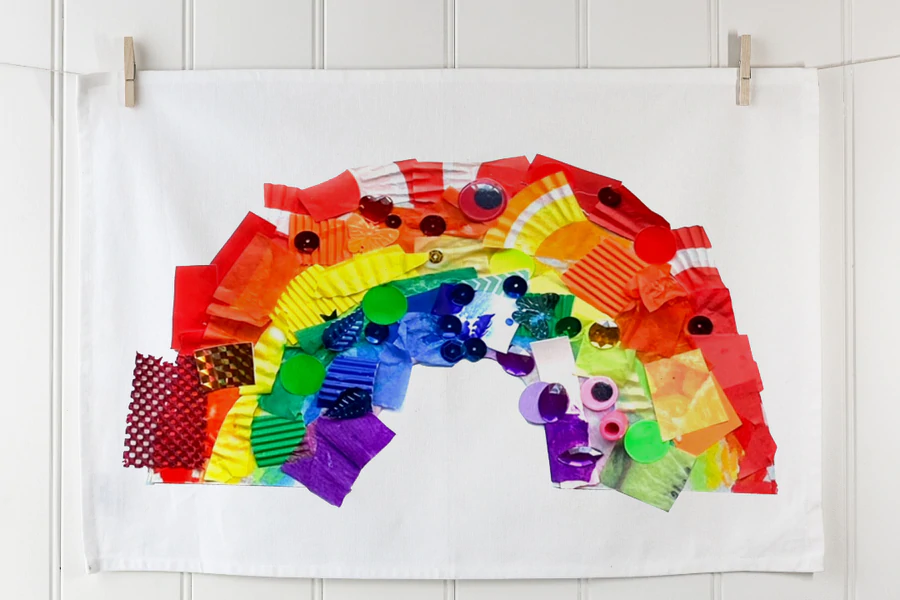 Creative fundraising ideas – rainbow collage made from colourful craft materials, printed on a tea towel and displayed on a white wall with pegs