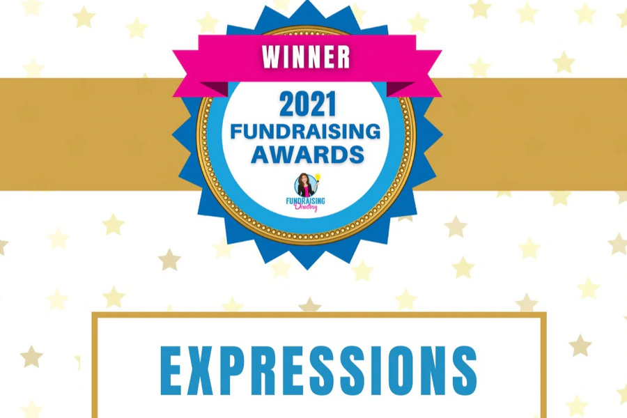 A celebratory graphic with a gold ribbon and starry background featuring a bold blue and gold award badge. At the center of the badge, it reads “2021 Fundraising Awards” with the “Fundraising Directory” logo underneath. A bright pink ribbon banner across the top of the badge says “WINNER”. Below the badge, inside a framed box, the word “EXPRESSIONS” is prominently displayed in blue capital letters.
