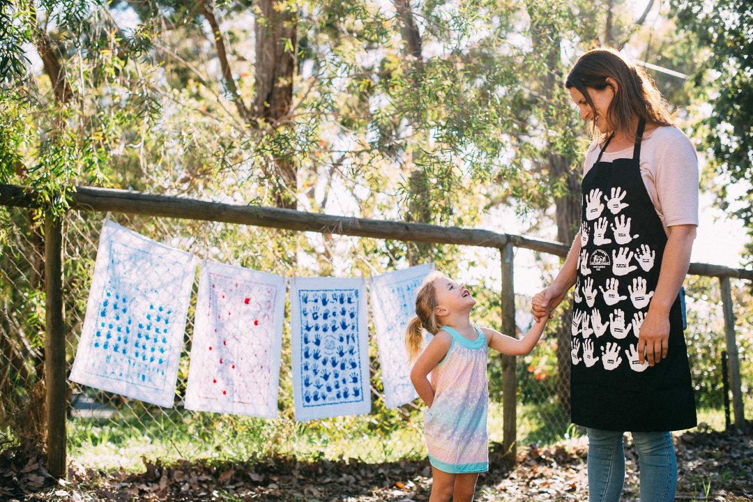 Mother and daughter holding hands in a sunlit backyard, standing beside a clothesline with colourful Expressions tea towel keepsakes printed with children's handprints.