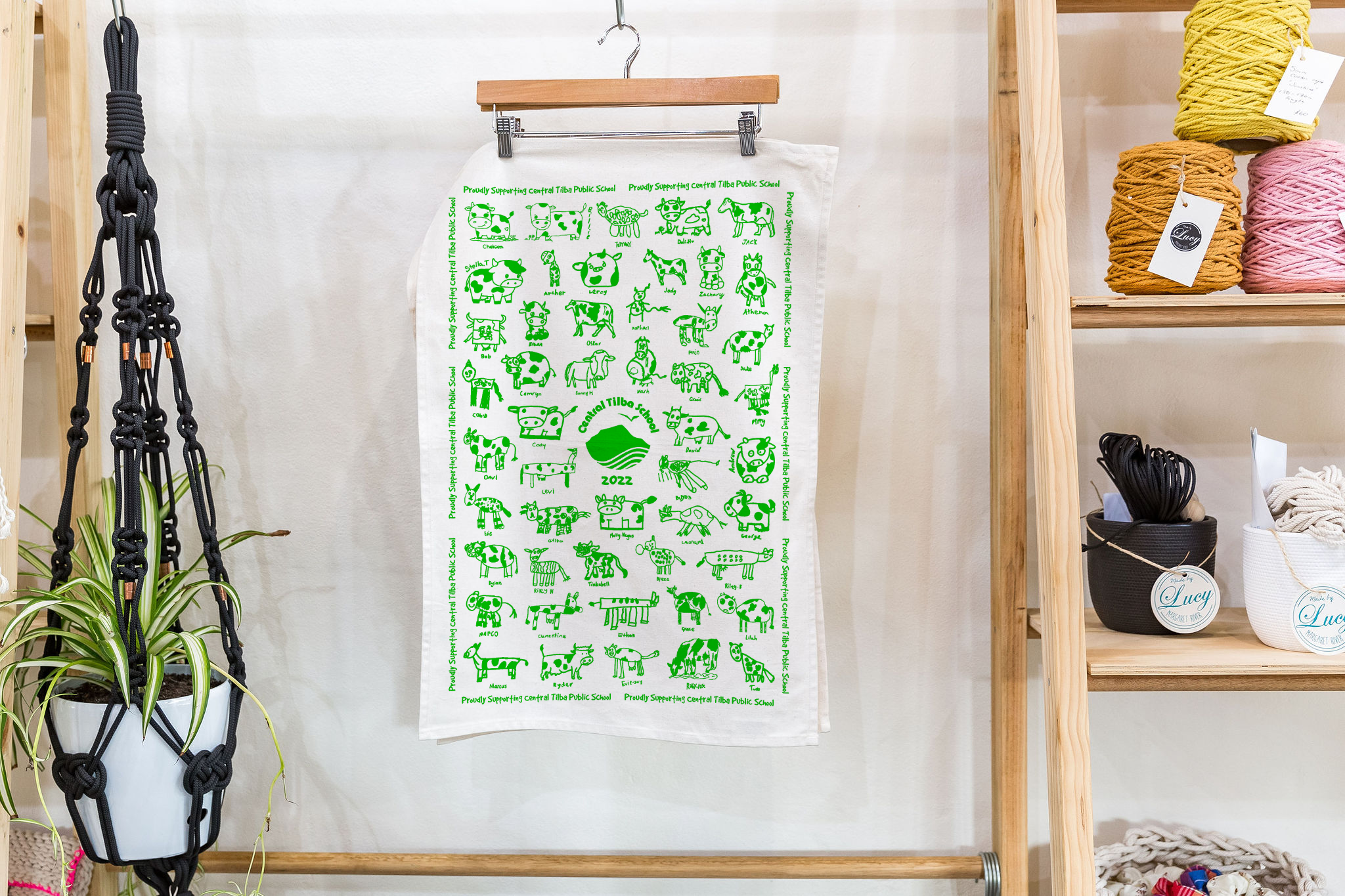 Custom tea towel hanging on display featuring green cow drawings by students from Central Tilba Public School, celebrating a rural-themed fundraising project.