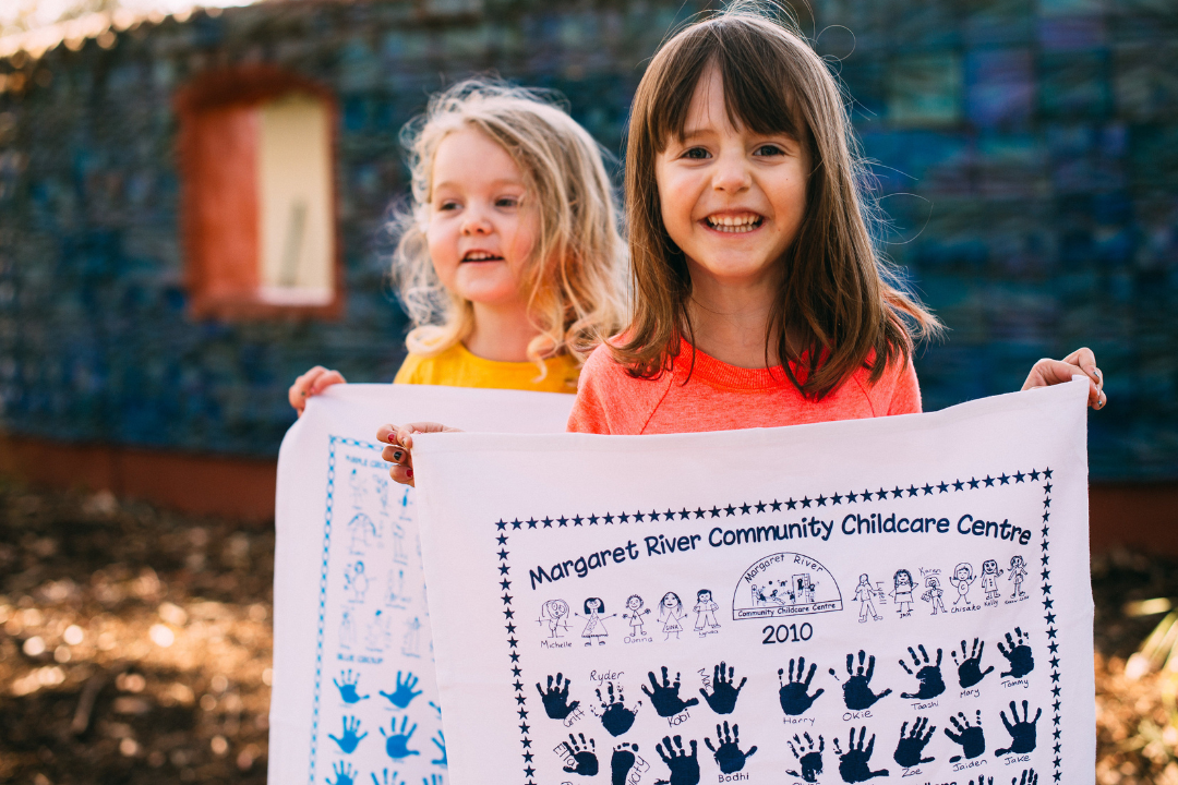 Two smiling young children proudly holding custom tea towels featuring handprints and drawings from Margaret River Community Childcare Centre — part of an Expressions Tea Towel fundraiser in Australia.