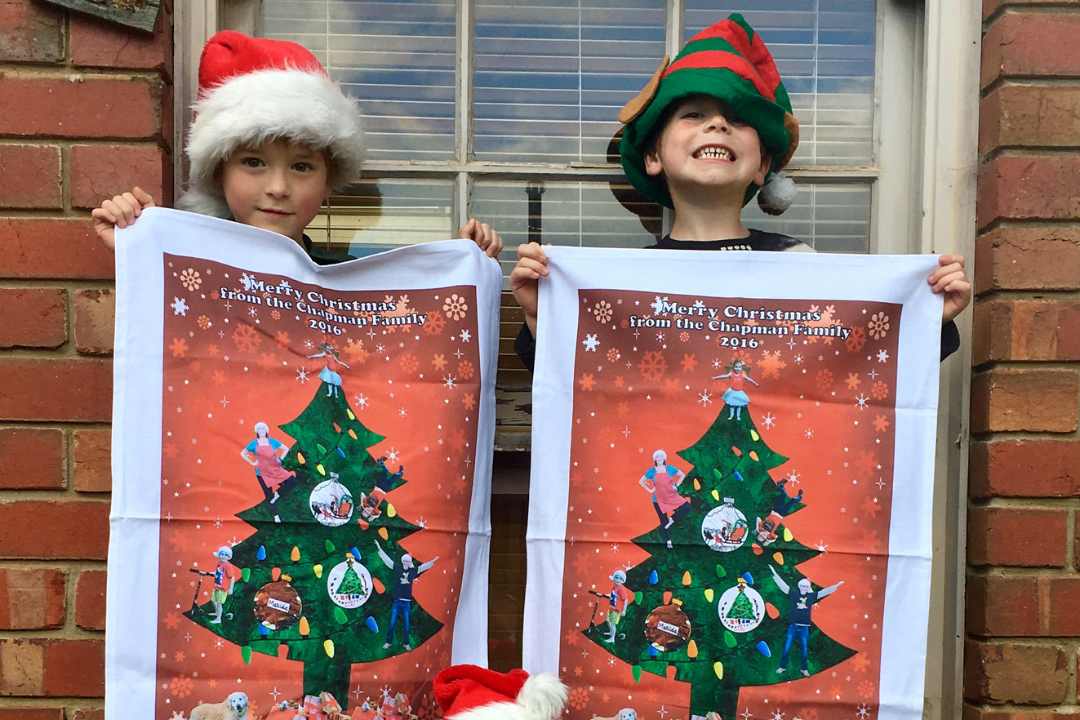 Two smiling children in Santa and elf hats proudly holding personalised Christmas tea towels with festive tree artwork and family greeting—Expressions Fundraising.