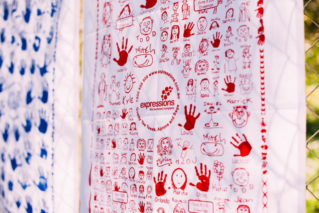 Close-up of an Expressions Tea Towel featuring children's hand-drawn portraits, names, and red handprints, showcasing a collaborative school fundraising design.