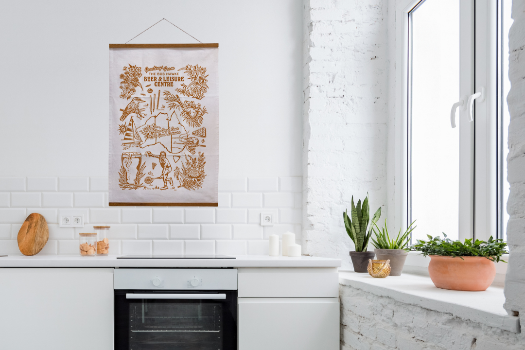 A warm white kitchen featuring a Bob Hawke Beer & Leisure Centre tea towel, styled with indoor plants and rustic ceramics.