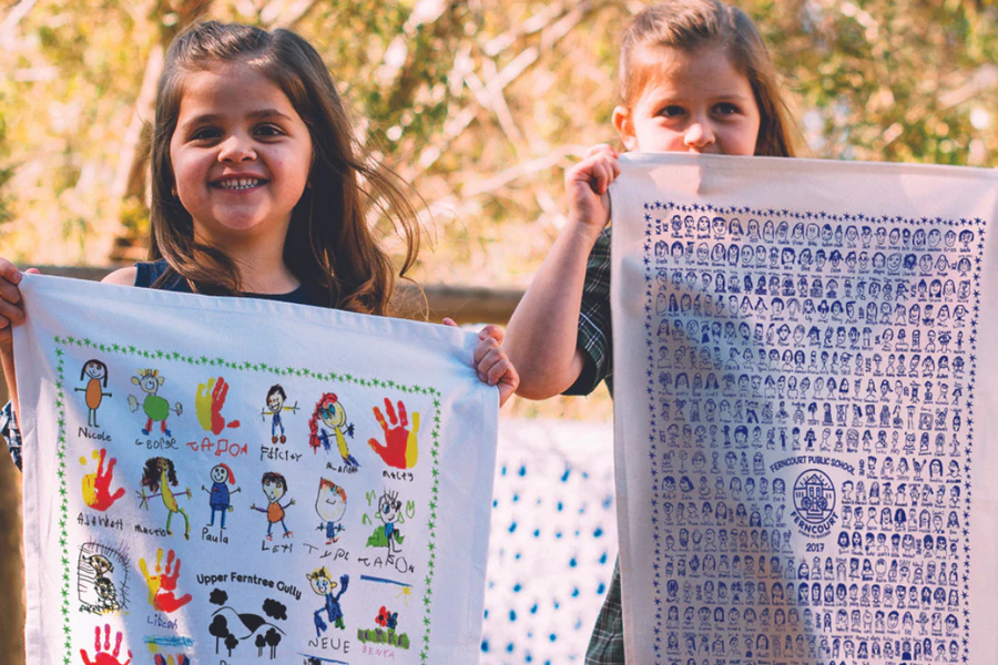 Two young schoolgirls proudly holding custom tea towels—one with colourful handprints and drawings, the other with class portraits—capturing school fundraiser memories.