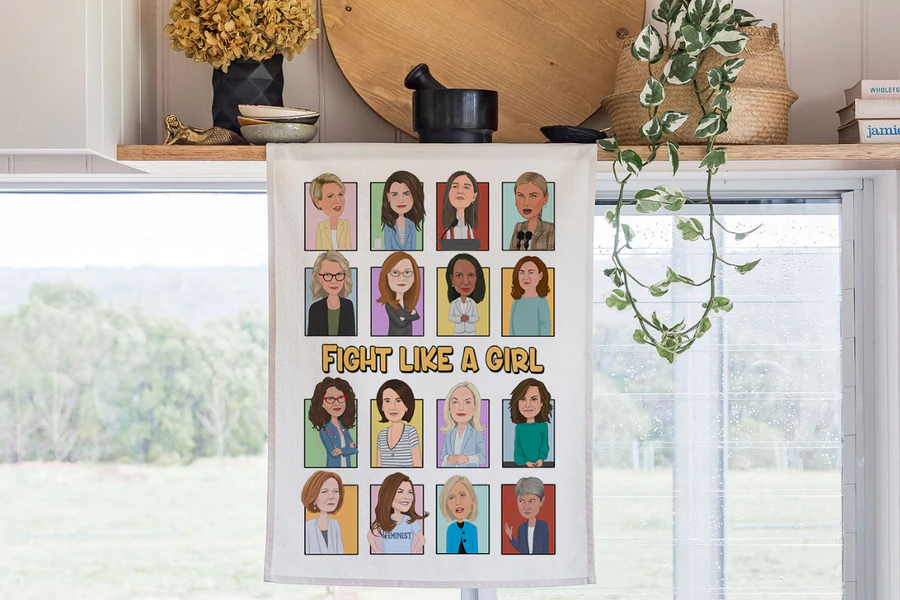 Fight Like a Girl tea towel hanging in a bright kitchen. The design features illustrated portraits of iconic Australian women with the bold text 'Fight Like a Girl' in the centre. Made from organic cotton by Expressions Australia.