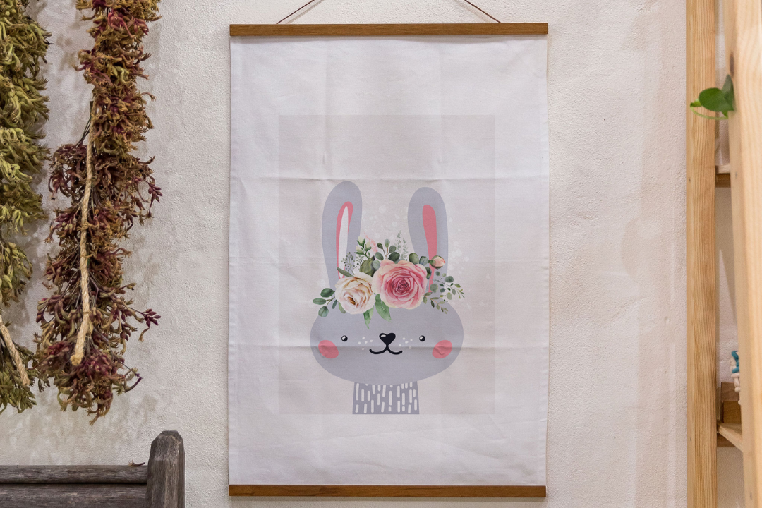 Illustration of a grey bunny with pink cheeks and inner ears, wearing a flower crown made of roses and greenery, printed on a white tea towel and displayed in a wooden hanger frame.