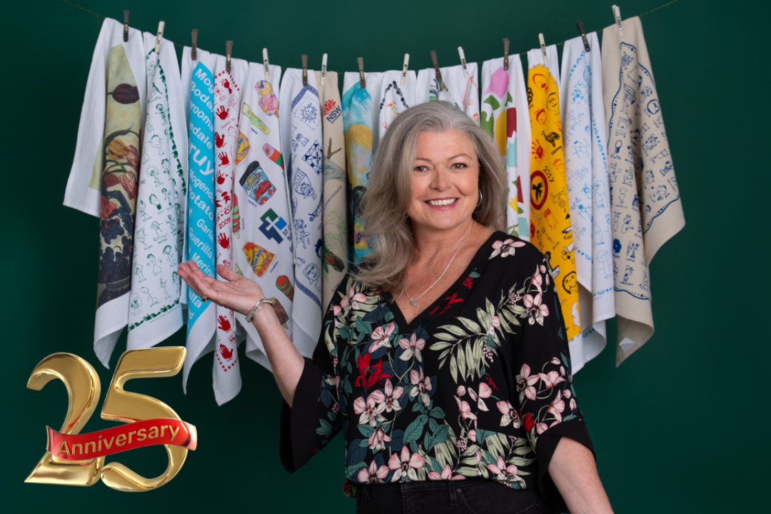 Founder of Expressions smiling in front of a colourful display of custom printed tea towels, celebrating 25 years of ethical fundraising in Australia.