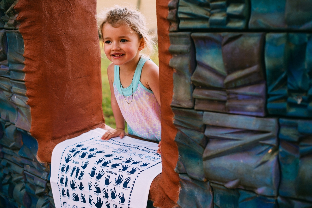 Young child proudly holding a custom printed school tea towel featuring handprints and drawings, created for a Community Childcare Centre fundraiser by Expressions Australia