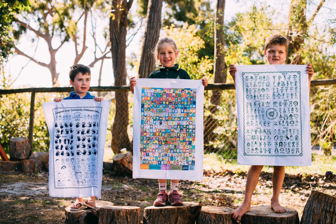 Three smiling school children proudly holding up custom tea towels featuring colourful self-portraits, handprints and classroom artwork showcasing creative school fundraising projects by Expressions Australia.