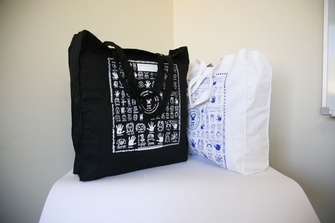 Two custom printed tote bags from Expressions Fundraising featuring children's hand-drawn artwork; one in black with white prints and one in white with blue prints, displayed on a white surface
