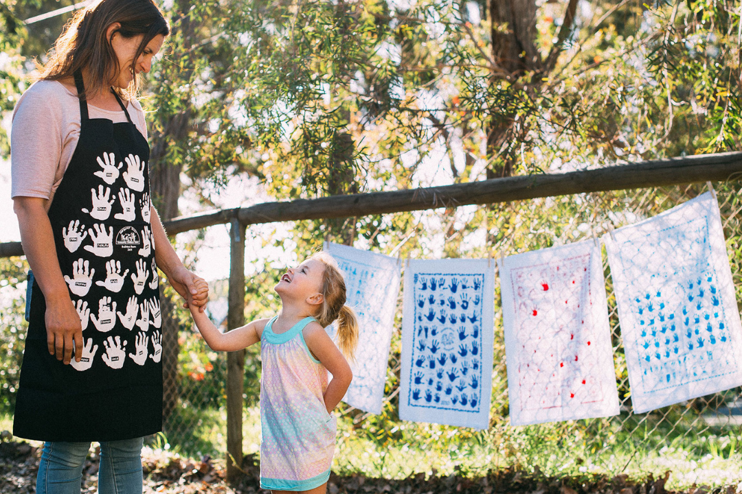Mother and daughter smiling at each other outdoors beside a fence decorated with colourful handprint tea towels.