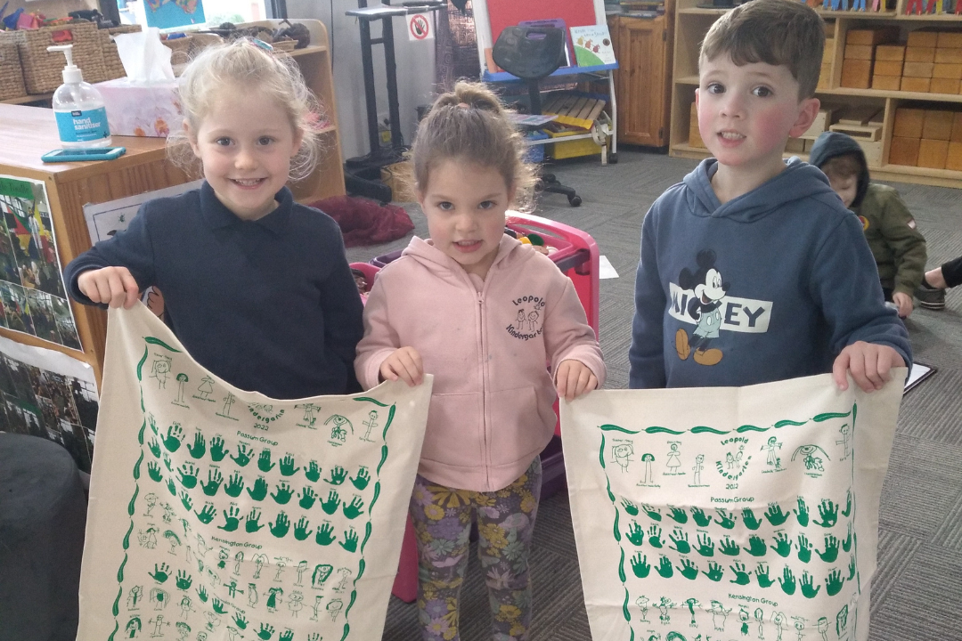 Three smiling kindergarten children proudly holding custom-printed tea towels featuring handprints and drawings at Leopold Kindergarten’s fundraising event.