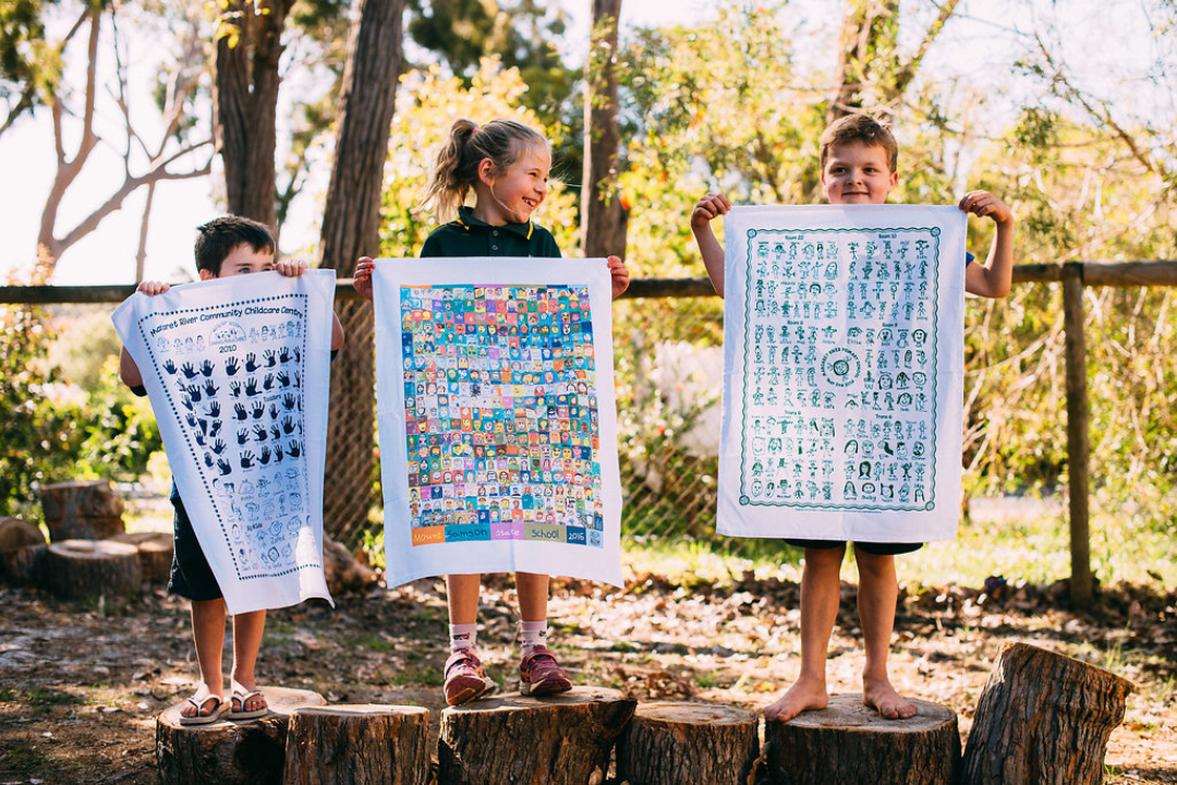 Three young children proudly holding colourful printed tea towels outdoors, showcasing their school artwork as part of a creative fundraising project by Expressions.