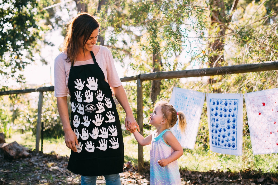 Child wearing a black school fundraiser apron printed with student self-portraits, standing on a wooden bridge at a school playground