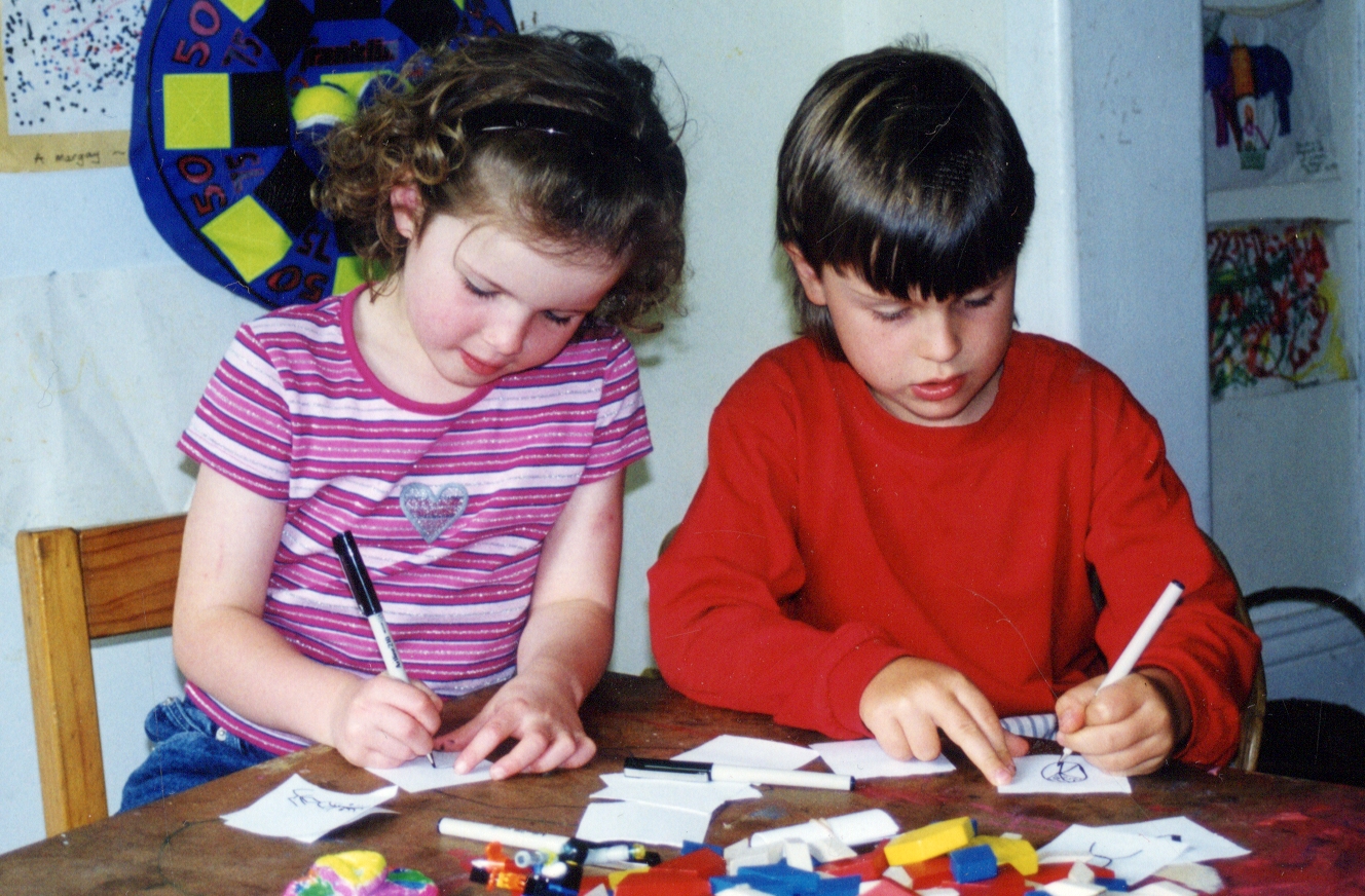 Two young children sit at a table drawing with markers on small pieces of paper, surrounded by colourful blocks and classroom artwork, creating designs for a school fundraiser project.